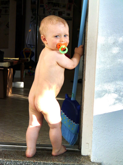 naked baby stands in a doorway and holds a toy broom